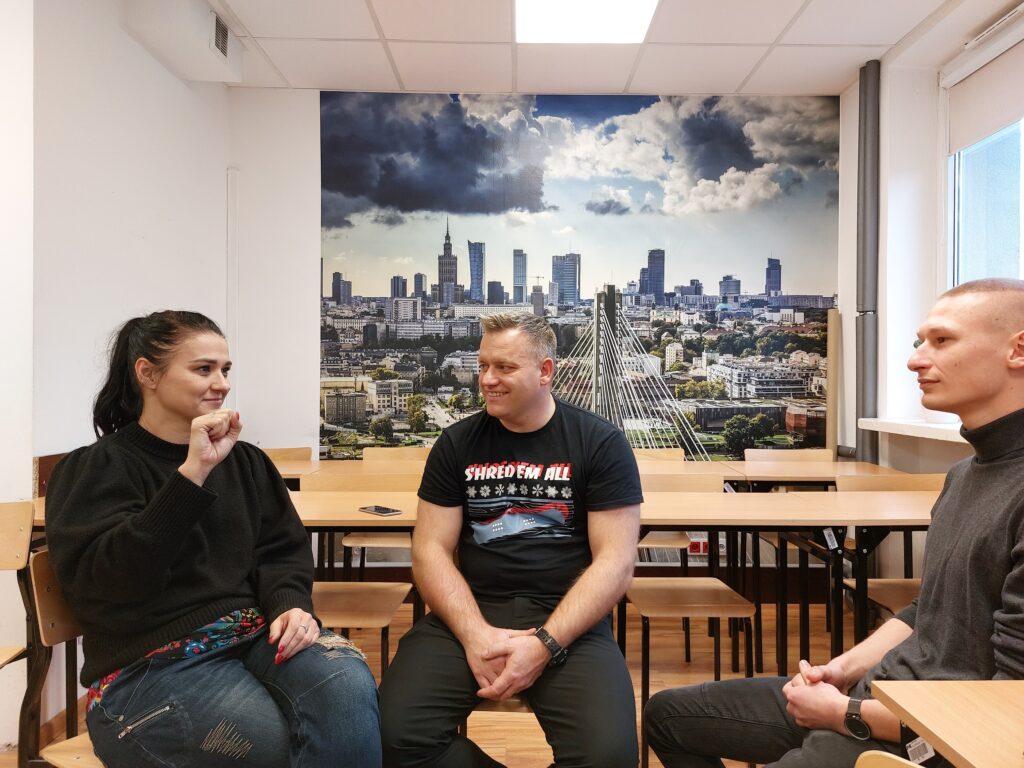 Trzy osoby na zajęciach z języka migowego, kobieta pokazuje znak litery "B" - zaciśniętą pięść